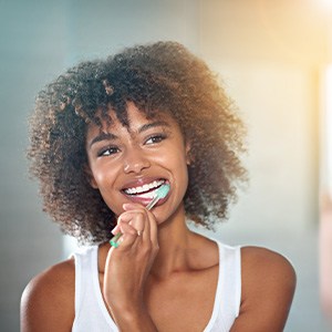 Woman in white tank top smiling into mirror while brushing her teeth