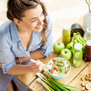 Woman in striped shirt leaning on wooden table eating salad next to fresh fruits and veggies